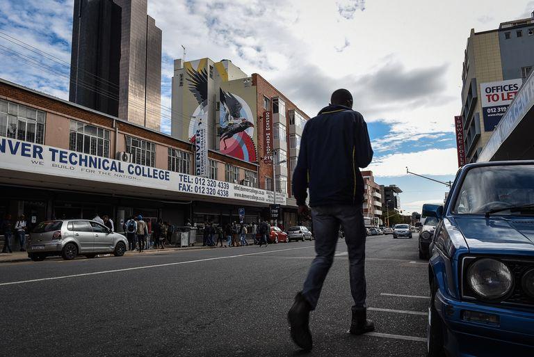 A man walks away down a city street lined with high-rise buildings and painted signage, cars parked at the kerb.