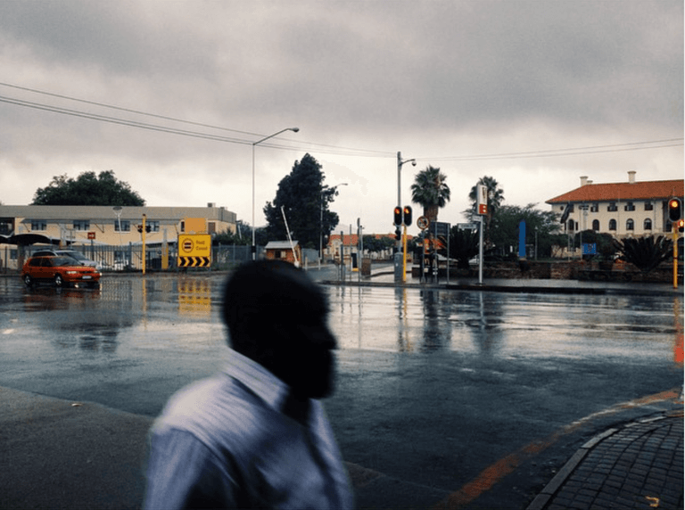 A man in a white shirt walks through a rain-soaked intersection at dusk, his head a dark silhouette against wet tar.