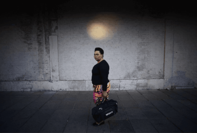 A woman in a black top and patterned skirt walks along a pavement carrying a sports bag, beneath a round pool of light on a dark wall.